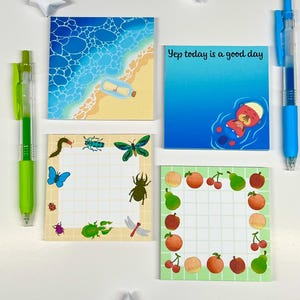 May include: Four square sticky notes with different designs. One depicts a beach scene with a message in a bottle. Another says "Yep today is a good day" with an otter. The others have insect and fruit illustrations. Two pens and paper stars are also visible.