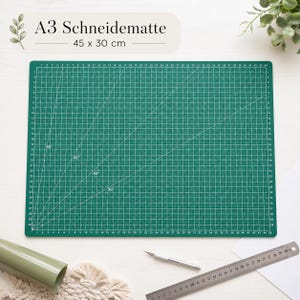 Könnte beinhalten: Eine grüne A3-Schneidematte mit weißen Rasterlinien und Winkelmarkierungen. Die Matte misst 45 x 30 cm und trägt oben den Text "A3 Schneidematte". Eine Rolle grünen Materials, ein Bastelmesser und ein Lineal sind ebenfalls zu sehen.