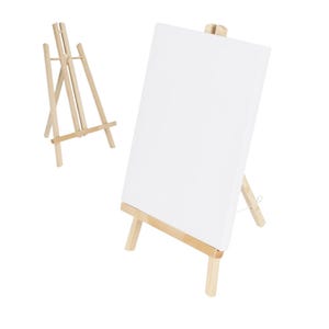 Könnte beinhalten: Zwei kleine Holzstaffeleien, eine davon hält eine leere weiße Leinwand. Die Staffeleien sind aus hellem Holz gefertigt und für die Aufnahme von Kunstwerken konzipiert. Die Leinwand ist rechteckig und bereit zum Malen oder Zeichnen.