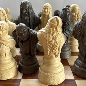 May include: A detailed chess set featuring character busts in shades of beige and dark brown. The chess pieces are arranged on a wooden checkered board, showcasing intricate details and varying textures.