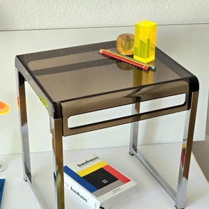 May include: A square side table with a dark translucent brown acrylic top and a chrome metal frame. On the table are two coloured pencils, a yellow acrylic block, and a small yellow cube. A book titled Bauhaus sits below the table.
