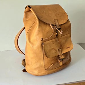 Könnte beinhalten: Ein beigefarbener Lederrucksack mit einer großen Klappe mit Schnallenverschluss, einem Hauptfach mit Kordelzug und einer Vordertasche mit Schnallenriemen. Der Rucksack verfügt über verstellbare Schultergurte und den eingeprägten Markennamen BREE.