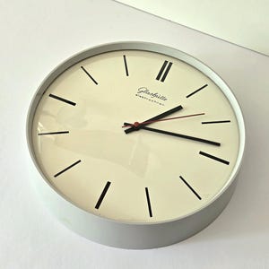 Könnte beinhalten: Eine runde Wanduhr mit hellgrauem Rahmen und cremefarbenem Zifferblatt. Die Uhr hat schwarze Stundenmarkierungen, schwarze Stunden- und Minutenzeiger sowie einen dünnen roten Sekundenzeiger. Der Schriftzug Glashütte elektrochron ist in Schwarz aufgedruckt.