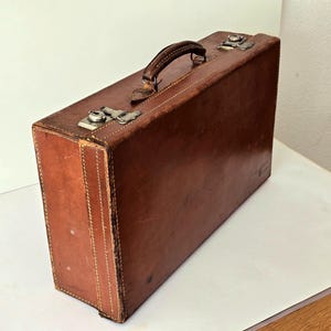 Könnte beinhalten: Ein rechteckiger Aktenkoffer aus braunem Leder mit einem Tragegriff an der Oberseite, sichtbaren Nähten an den Kanten und zwei silberfarbenen Metallverschlüssen. Die Lederoberfläche weist natürliche Abnutzungserscheinungen, Kratzer und Farbvariationen auf.