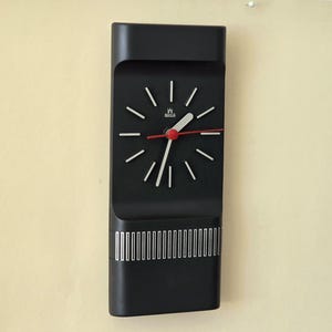 Könnte beinhalten: Rechteckige schwarze Wanduhr mit weißen Stundenmarkierungen und Zeigern sowie einem roten Sekundenzeiger. Die Uhr hat ein minimalistisches Design mit silbernen Streifen am unteren Rand. Sie hängt an einer hellbeigen Wand.