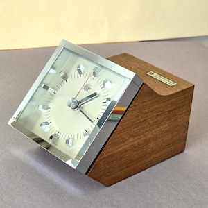 Könnte beinhalten: Eine Vintage-Junghans-Uhr mit silberfarbenem Zifferblatt und Zeigern, eingebettet in ein rechteckiges, abgewinkeltes, klares Glasgehäuse. Die Uhr ist auf einem Holzsockel mit natürlicher Holzmaserung montiert.