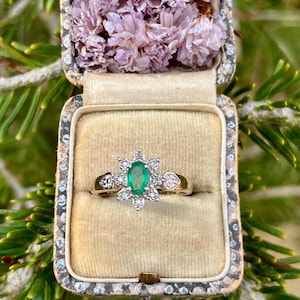 May include: A gold ring with an oval emerald and diamond cluster, presented in a vintage box. The box is lined with beige fabric and filled with dried purple flowers. The ring's design includes heart-shaped accents.