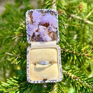 May include: An antique engagement ring displayed in an open, decorative box. The ring features a gold band and a central square stone. The box is lined with velvet and filled with dried purple flowers. The background is a close-up of green fir tree needles.