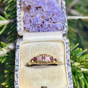 May include: A gold ring with three red gemstones and smaller white gemstones set in a textured band. The ring is in a small, velvet-lined box.