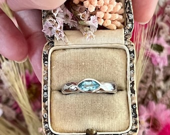 Puede incluir: Un anillo de tono plateado con una piedra preciosa azul claro de corte marquesa, engastada en una banda de metal liso. El anillo se presenta dentro de un pequeño joyero rectangular forrado de terciopelo beige con borde estampado.