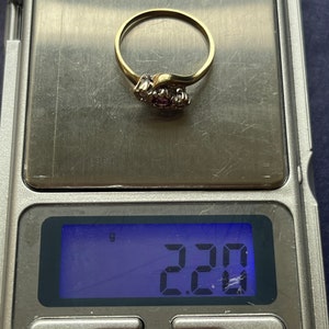 以下が含まれることがあります： 小さな丸い宝石のクラスターが付いたゴールドリング。 リングはデジタルスケール上にあり、2.20グラムの重量が表示されています。 リングは繊細で湾曲したデザインです。