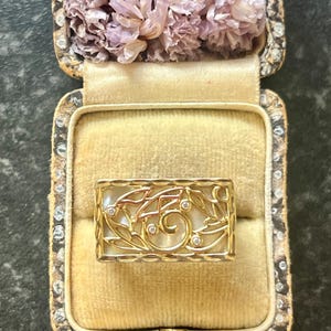 May include: A rectangular gold ring with a mother-of-pearl inlay and a filigree design featuring leaves and small diamonds. The ring is displayed in an antique jewellery box with dried flowers.