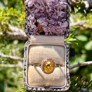 May include: A vintage gold ring with a large, oval, yellow gemstone, displayed in an open, antique jewelry box filled with dried lavender flowers. The ring is set against a beige velvet interior. The box is framed with a decorative border.