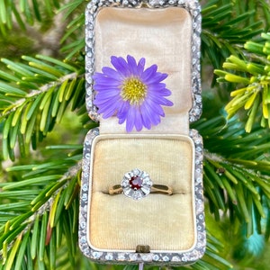 May include: A red gemstone ring with a white gold band and small diamonds set in a flower design. The ring is in a small, cream-coloured box with a purple flower on top.