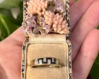 May include: A gold band ring featuring a row of alternating dark blue rectangular stones and small clear stones, presented in a small, rectangular, patterned box with a beige velvet interior and dried pink and purple flowers.