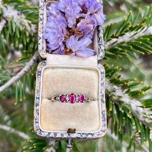 May include: A gold ring featuring three oval-cut red rubies set in a row, accented by small clear stones on each side. The ring is displayed inside a vintage-style rectangular jewelry box lined with beige fabric and topped with purple flowers.