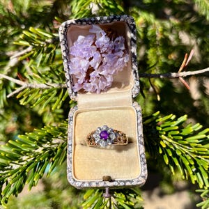 Puede incluir: Un anillo de oro con una piedra preciosa púrpura y un diseño floral. El anillo está en una pequeña caja blanca con un diseño floral en la tapa.