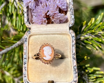 Op de afbeelding: Een gouden ring met camee in een open vintage sieradendoosje gevuld met gedroogde lavendelbloemen. De ring heeft een vrouwenprofiel in een ovale lijst. Het doosje is gevoerd met beige fluweel en heeft een decoratief, sierlijk ontwerp.