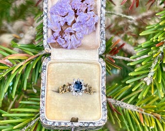 May include: A gold ring featuring an oval blue sapphire center stone surrounded by a halo of small clear diamonds, set within a vintage-style jewelry box lined with beige velvet and decorated with dried purple flowers inside the lid.