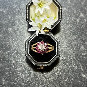 May include: A gold ring with a large pink gemstone surrounded by smaller white gemstones in a black velvet ring box with white flowers.