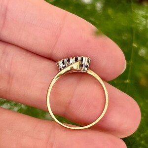 以下が含まれることがあります： 小さな暗い宝石のクラスターが付いたゴールドリング。 リングのバンドは細くて円形で、宝石のセッティングは対照的なシルバーカラーです。 リングは人の手で持たれており、緑色の背景があります。