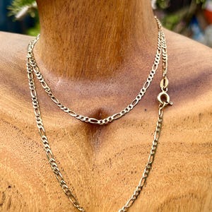 May include: A gold-toned figaro chain necklace featuring a series of flattened circular links interspersed with elongated oval links. The necklace is secured with a round spring ring clasp, showcasing a polished metallic finish and a classic link design.