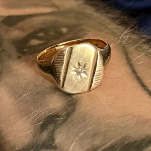 Pode incluir: Um anel de sinete dourado com uma face retangular com um design de estrela e linhas verticais. O anel tem um acabamento polido e uma banda arredondada. Uma peça de joalharia clássica.