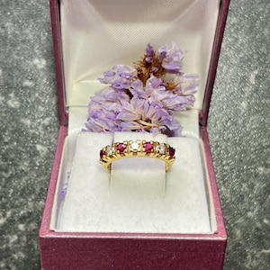 May include: A gold band ring featuring an alternating row of round-cut red rubies and clear white diamonds. The ring is displayed inside a purple hinged jewelry box with a white satin interior lining and a small cluster of dried purple flowers.