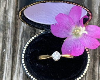 May include: A gold ring with a single round diamond in a black ring box with a purple flower on top.