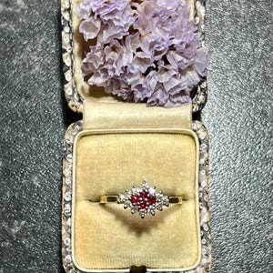 以下が含まれることがあります： 赤いルビーと白いダイヤモンドがクラスターデザインにセットされたゴールドリング。リングは、紫色のドライフラワーが入った小さなベルベットボックスに入っています。