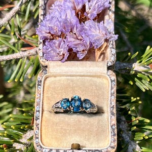 May include: A silver ring featuring three oval-cut blue gemstones set in a row, displayed inside a vintage-style rectangular jewellery box lined with beige velvet and decorated with dried purple flowers.