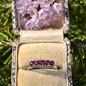 May include: A silver ring set with five vivid red gemstones, presented in an antique jewellery box with a beige velvet lining. The open box displays a cluster of delicate, pale purple flowers. The box has a decorative, patterned border.