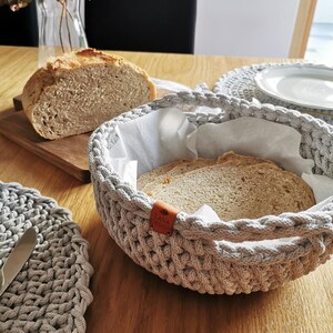Op de afbeelding: Een grijze broodmand van geweven materiaal met gesneden brood, gevoerd met wit papier. Een brood zit op een houten plank. Een bijpassende placemat en een vaas met bloemen staan ook op de houten tafel.