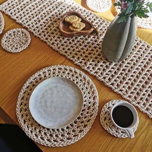 Może przedstawiać: Beżowy szydełkowy zestaw bieżnika, podkładek i podkładek na drewnianym stole. Na stole znajduje się również ceramiczny talerz, filiżanka kawy i drewniana miska z ciasteczkami. W tle wazon z różowymi kwiatami.