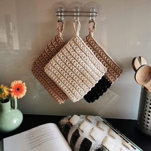 Può includere: Tre presine quadrate all'uncinetto in beige, beige chiaro e nero sono appese a un supporto metallico. Le presine hanno un anello per appenderle. Un vaso verde con fiori arancioni e gialli è a sinistra. Un libro di cucina aperto è in primo piano.
