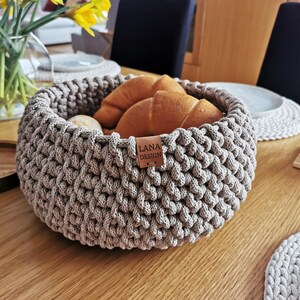 Op de afbeelding: Een ronde, grijze gehaakte mand gevuld met goudbruine croissants en broodjes. De mand heeft een dikke, getextureerde weefsel en een klein leren label met de tekst "LANA DESIGN". De mand staat op een houten tafel.