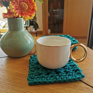 Puede incluir: Una taza de cerámica blanca con asa dorada descansa sobre un posavasos de ganchillo turquesa. Un jarrón verde con flores naranjas y amarillas está en el fondo, todo sobre una mesa de madera. La escena está en una habitación bien iluminada.