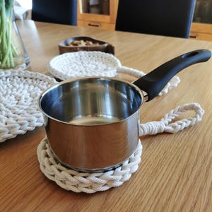 Może przedstawiać: Garnek ze stali nierdzewnej z czarnym uchwytem spoczywa na kremowej, ręcznie robionej podkładce. Pasująca podkładka i mała drewniana miska również znajdują się na drewnianym stole. Podkładki mają średnicę około 15 cm.