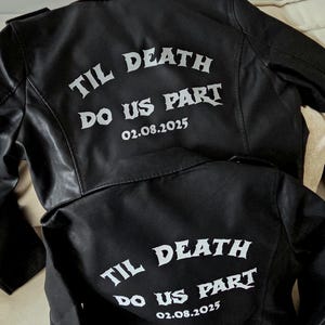 Könnte beinhalten: Zwei schwarze Lederjacken mit dem weißen Aufdruck "TIL DEATH DO US PART 02.08.2025" auf dem Rücken. Die Jacken werden auf einer neutralfarbenen Oberfläche präsentiert. Die Jacken haben ein klassisches Bikerjacken-Design.