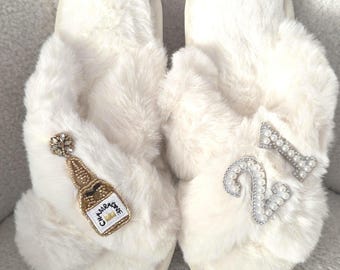 May include: A pair of white, fluffy slippers. One slipper features a gold and pearl champagne bottle design, the other has a pearl-studded number "2" and a bow. The slippers are designed for indoor wear.