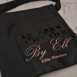May include: Black leather makeup artist's apron with multiple pockets and a strap. The apron has the text "By Ell Ellie Hawkes" printed in pink.