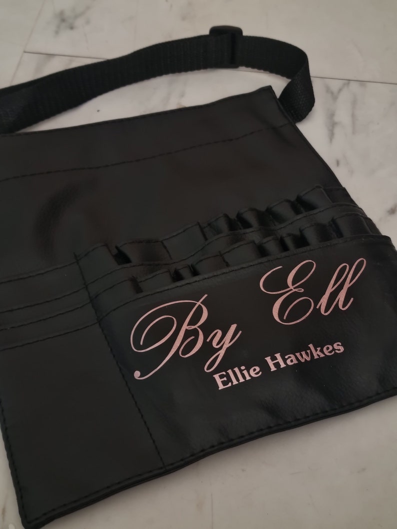 May include: Black leather makeup artist's apron with multiple pockets and a strap. The apron has the text "By Ell Ellie Hawkes" printed in pink.