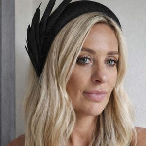 May include: A black headband featuring a cluster of black feathers on the left side. The headband is worn by a person with long, wavy blonde hair. The background is a plain, light-coloured wall. Close-up shot.