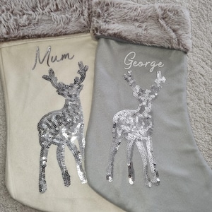May include: Two Christmas stockings with a reindeer design. One stocking is white with a gray fur cuff and the name "Mum" embroidered on it. The other stocking is gray with a gray fur cuff and the name "George" embroidered on it.