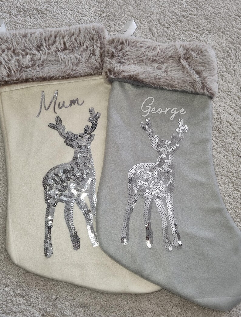 May include: Two Christmas stockings with a reindeer design. One stocking is white with a gray fur cuff and the name "Mum" embroidered on it. The other stocking is gray with a gray fur cuff and the name "George" embroidered on it.