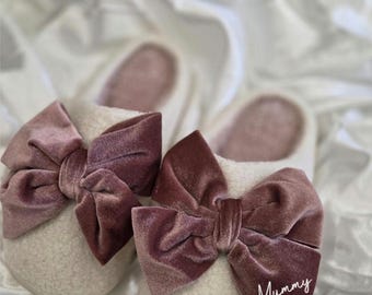 Può includere: Un paio di pantofole color crema con grandi fiocchi di velluto malva. La parola "Mummy" è scritta in corsivo su uno dei fiocchi. Le pantofole hanno un aspetto morbido e soffice, che suggerisce comfort e calore.
