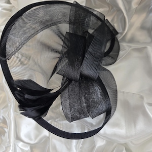 May include: A black fascinator hat with a mesh design, a large bow, and feathers. The hat has a circular base and is made of a sheer, lightweight material. The bow is made of a similar material and is positioned on the side of the hat.