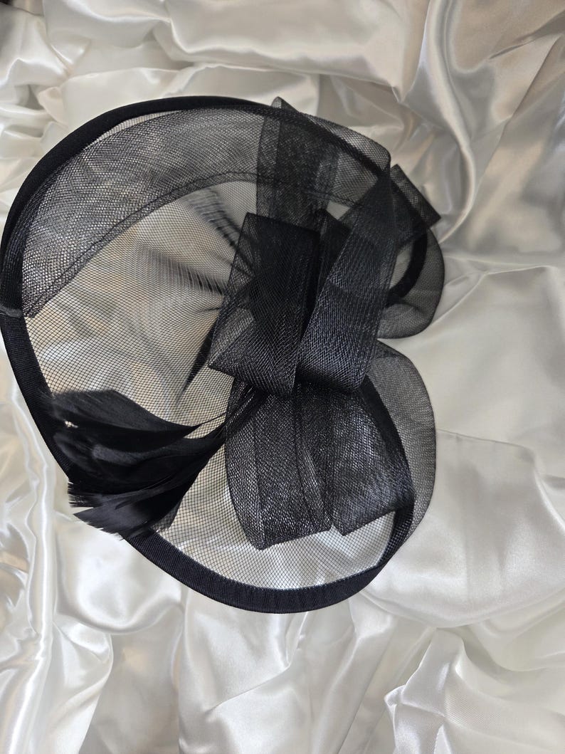 May include: A black fascinator hat with a mesh design, a large bow, and feathers. The hat has a circular base and is made of a sheer, lightweight material. The bow is made of a similar material and is positioned on the side of the hat.