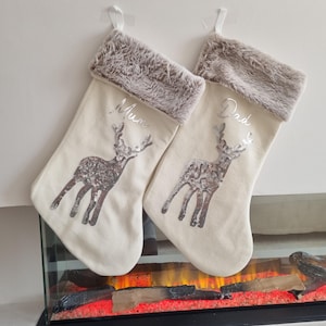 May include: Two cream-colored Christmas stockings with a reindeer design and silver sequins. The stockings have a brown faux fur cuff and are personalized with the names "Mum" and "Dad".