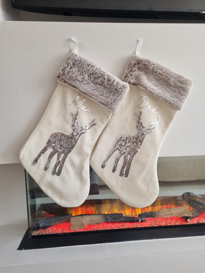 May include: Two cream-colored Christmas stockings with a reindeer design and silver sequins. The stockings have a brown faux fur cuff and are personalized with the names "Mum" and "Dad".